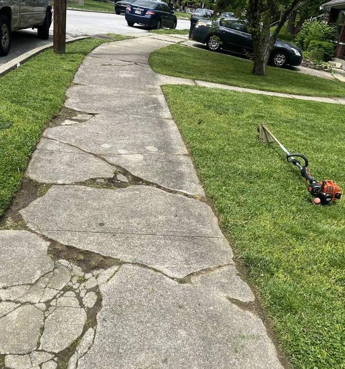 A string trimmer lying on a cracked concrete walk beside a freshly cut and edged lawn.
